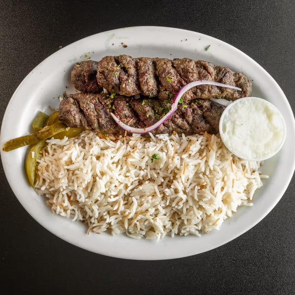 Two grilled meat skewers with rice, pickled peppers, and a side of white sauce on a plate.