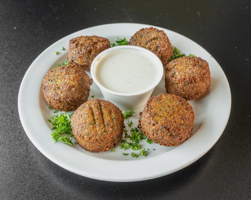 Six golden-brown falafel balls arranged around a small cup of white dipping sauce on a white plate.
