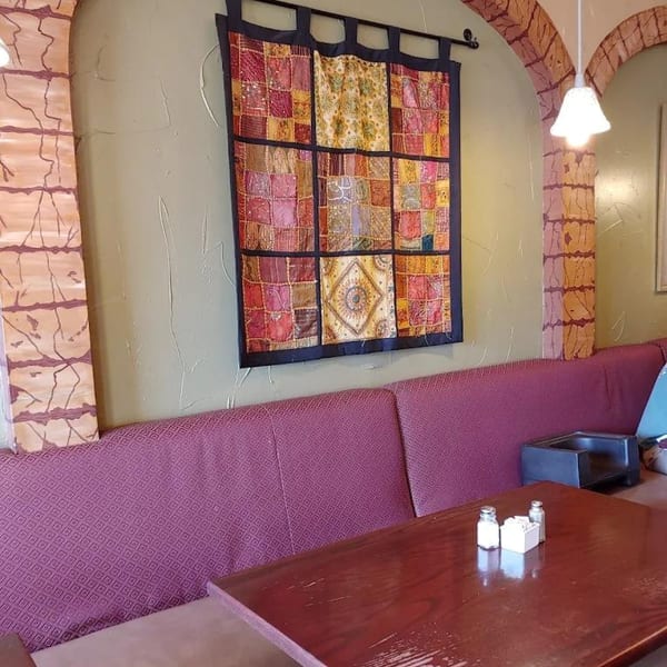 Restaurant interior with a wooden table, purple booth seating, and a colorful tapestry on a green wall.