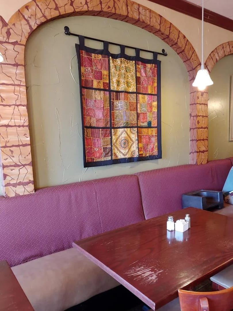 Restaurant interior with a wooden table, purple booth seating, and a colorful tapestry on a green wall.