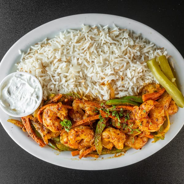 Shrimp and vegetable stir-fry with rice, a side of white sauce, and pickles on a white plate.
