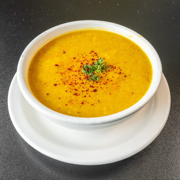 A bowl of yellow lentil soup, garnished with red pepper flakes and parsley, on a white plate.