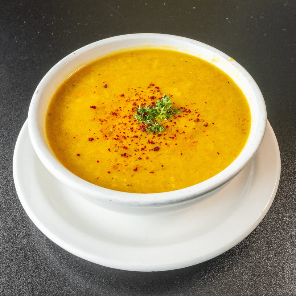 A bowl of yellow lentil soup, garnished with red pepper flakes and parsley, on a white plate.