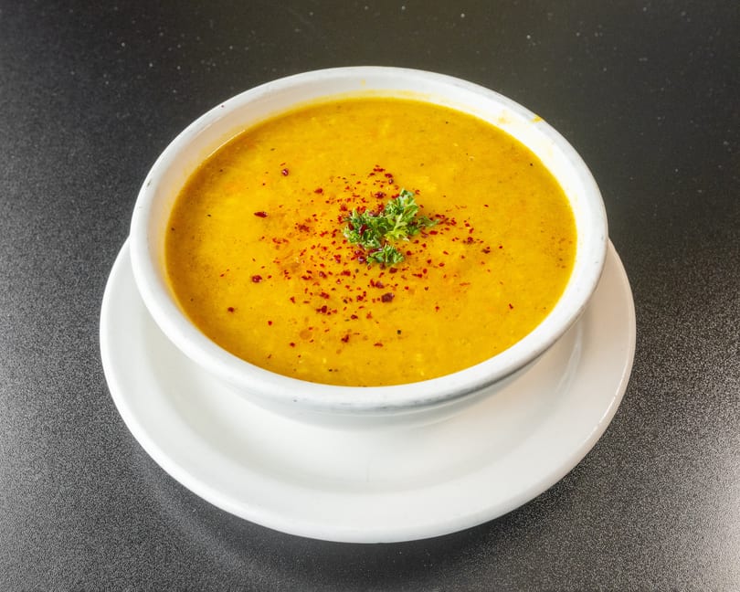 A bowl of yellow lentil soup, garnished with red pepper flakes and parsley, on a white plate.