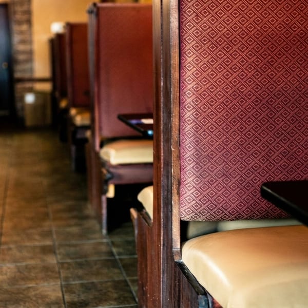 A row of empty restaurant booths with red patterned backs and tan seats, leading to a dark doorway.