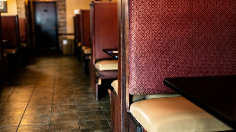 A row of empty restaurant booths with red patterned backs and tan seats, leading to a dark doorway.